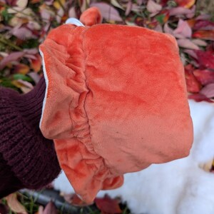 May include: A plush, orange fox-shaped hat with a white interior and a small, rounded ear detail. The hat is made of a soft, velvety material and has a gathered, elasticized edge. The hat is held up by a person wearing a dark maroon sweater.