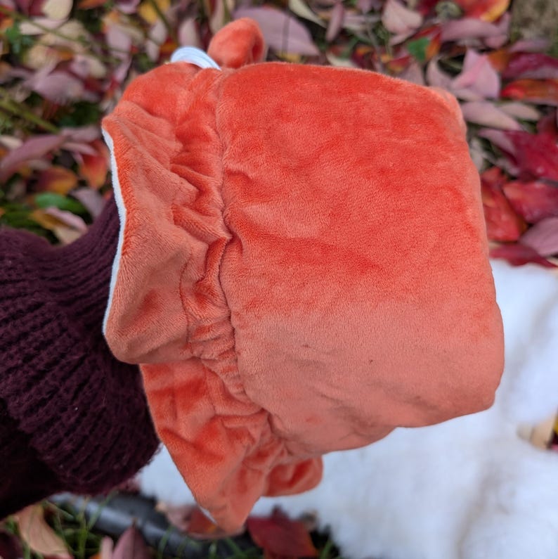 May include: A plush, orange fox-shaped hat with a white interior and a small, rounded ear detail. The hat is made of a soft, velvety material and has a gathered, elasticized edge. The hat is held up by a person wearing a dark maroon sweater.