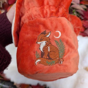 May include: An orange velvet drawstring bag featuring an embroidered fox design. The fox is brown with white accents, sitting peacefully with closed eyes. A crescent moon and foliage are also embroidered on the bag.