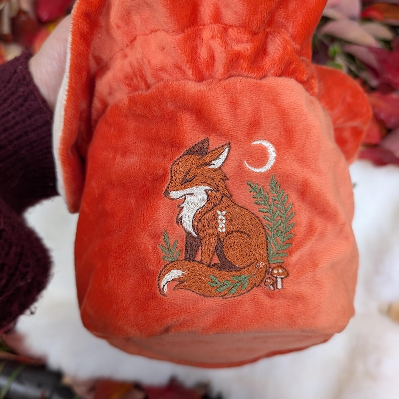 May include: An orange velvet drawstring bag featuring an embroidered fox design. The fox is brown with white accents, sitting peacefully with closed eyes. A crescent moon and foliage are also embroidered on the bag.