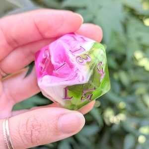 May include: A large, multi-sided die with a pink and green marbled design. The die has purple numbers and is held in a hand. The pink section has a white swirl pattern. The green section is translucent.