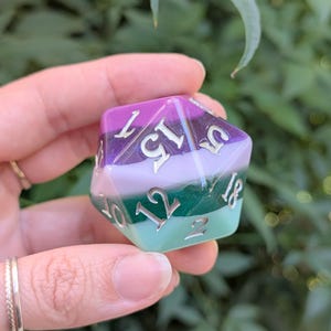 以下が含まれることがあります： 銀色の数字が入った、多色のD20サイコロ。サイコロは紫、ラベンダー、緑、アクアのグラデーションになっています。サイコロは手で持たれており、背景はぼやけた緑色です。
