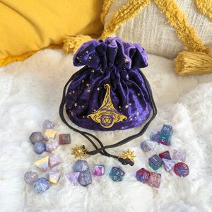May include: Purple velvet dice bag with gold embroidery of a wizard's hat and stars. The bag is open and spilling out a variety of colorful dice with different numbers and symbols.
