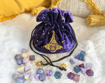 May include: Purple velvet dice bag with gold embroidery of a wizard's hat and stars. The bag is open and spilling out a variety of colorful dice with different numbers and symbols.