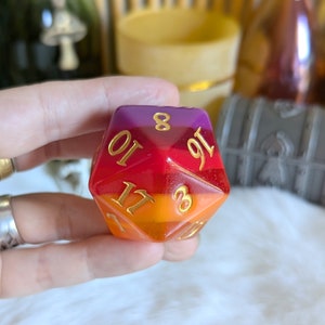 Puede incluir: Un dado d20 de resina transparente multicolor con números dorados. El dado está formado por capas de resina morada, roja y naranja. Los números en el dado son 3, 8, 10, 11, 16 y 19.