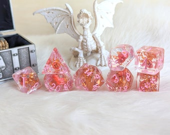 May include: A set of seven pink resin dice with embedded gold flakes and pink floral accents. The dice are in various geometric shapes, including a d4, d6, d8, d10, d12, d20, and a cube. A white dragon figurine is in the background.
