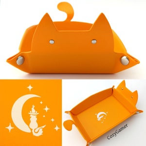 May include: Orange cat-shaped dice tray with a white silhouette of a cat sitting on a crescent moon with stars. The tray is made of felt and has a felt lining. The tray is designed to hold dice and other gaming accessories.