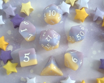 May include: A collection of pastel-colored polyhedral dice for tabletop games, including a D4, D6, D8, D10, D12, and D20. The dice are in shades of yellow, lavender, and white. Origami stars in yellow, purple, and white surround the dice.