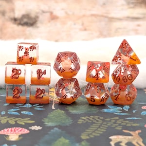 May include: A set of clear resin dice with orange accents and embedded fox designs. The dice include various shapes, such as cubes, d20s, and d4s, with numbers in a dark orange color. The dice are designed for tabletop gaming.