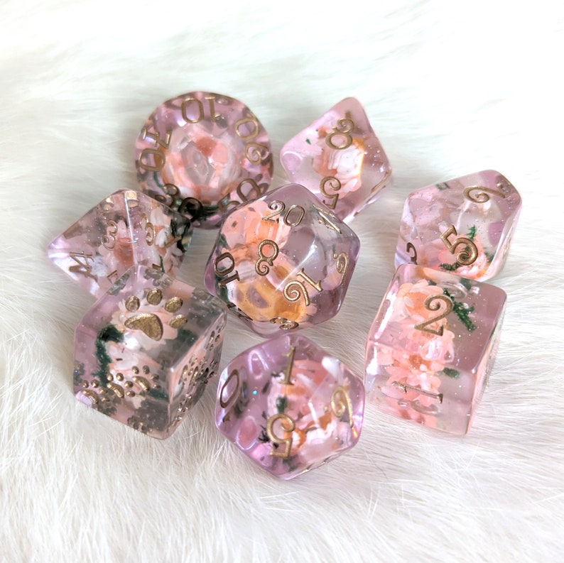 May include: A set of seven pink and clear dice with gold numbers and floral accents. The dice are arranged on a white fuzzy surface.
