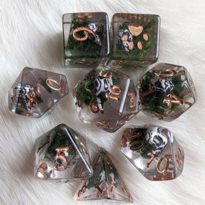 May include: A set of clear resin dice with copper-colored numbers. Each die features a miniature Dalmatian dog and green moss. The dice are in various shapes, including a cube, a d20, and a d4, perfect for tabletop gaming.