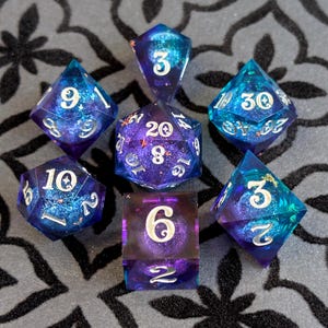 May include: Set of seven polyhedral dice for tabletop games. The dice are a mix of blue and purple with white numbering. The dice are made of resin and have a galaxy-like appearance. The dice are arranged in a circular pattern.