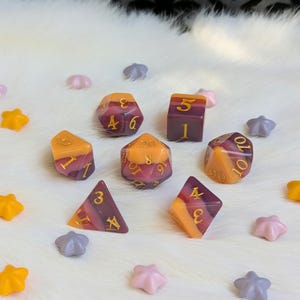May include: Set of seven dice with a purple and orange swirl pattern. The dice are numbered 1 through 6 and 10. The dice are on a white fluffy surface.