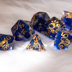 May include: A set of polyhedral dice in various shapes, featuring a deep blue hue with gold-colored numbering and decorative accents. The dice are made of a translucent material, with a glittery effect.