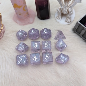 May include: A set of 10 purple dice with white numbers and silver glitter. The dice are a d4, d6, d8, d10, d12, and a d20. The dice are on a white fluffy surface.