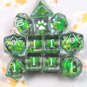 May include: A set of clear resin dice with green turtle and paw print designs. The dice include various shapes, such as a d4, d6, d8, d10, d12, and d20. The numbers are in white. These dice are ideal for tabletop games.