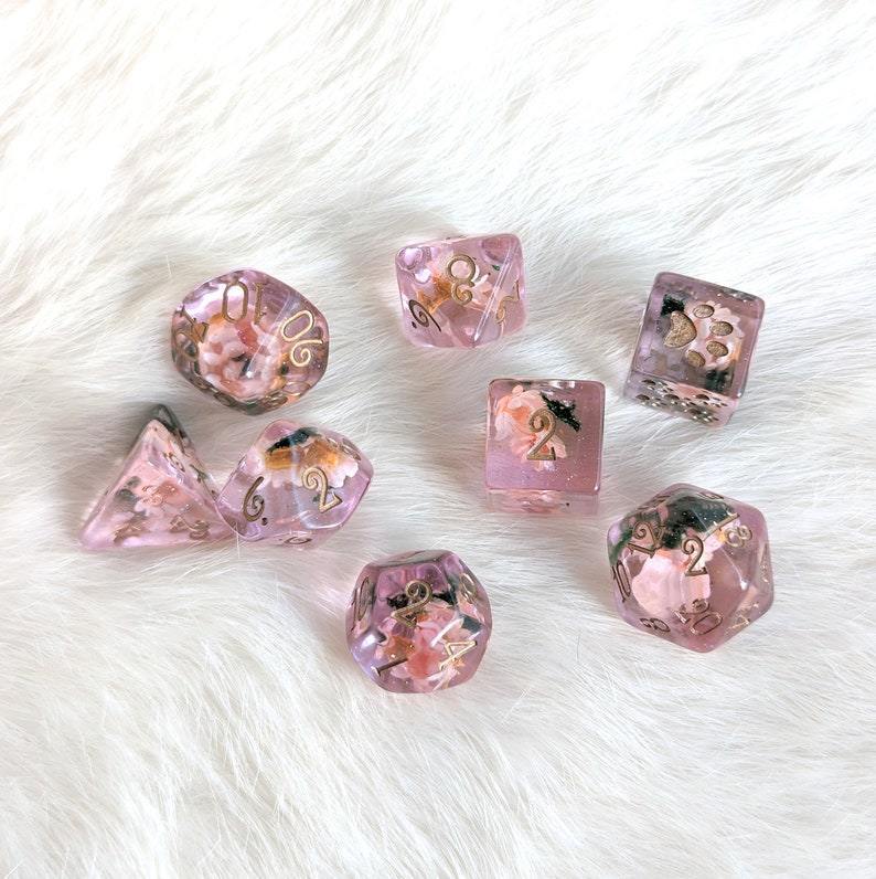 May include: Set of eight pink and gold dice with floral and paw print designs. The dice are translucent and have gold numbers.