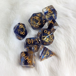May include: A set of seven blue and gold dice with gold glitter and various numbers on each face. The dice are on a white fluffy surface.