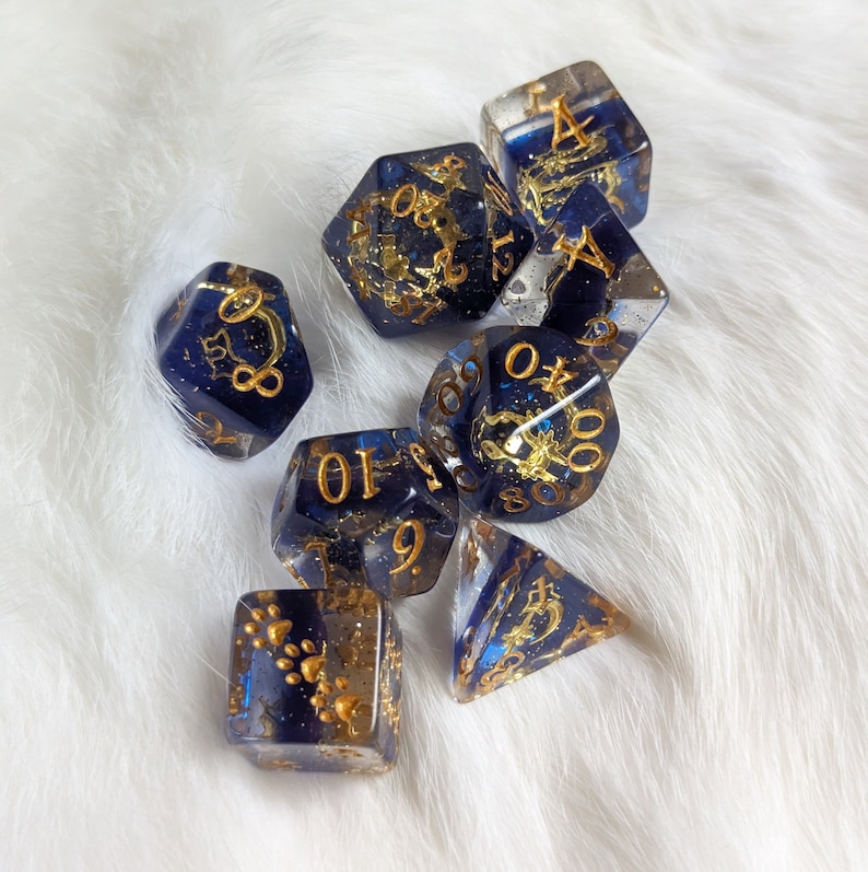 May include: A set of seven blue and gold dice with gold glitter and various numbers on each face. The dice are on a white fluffy surface.