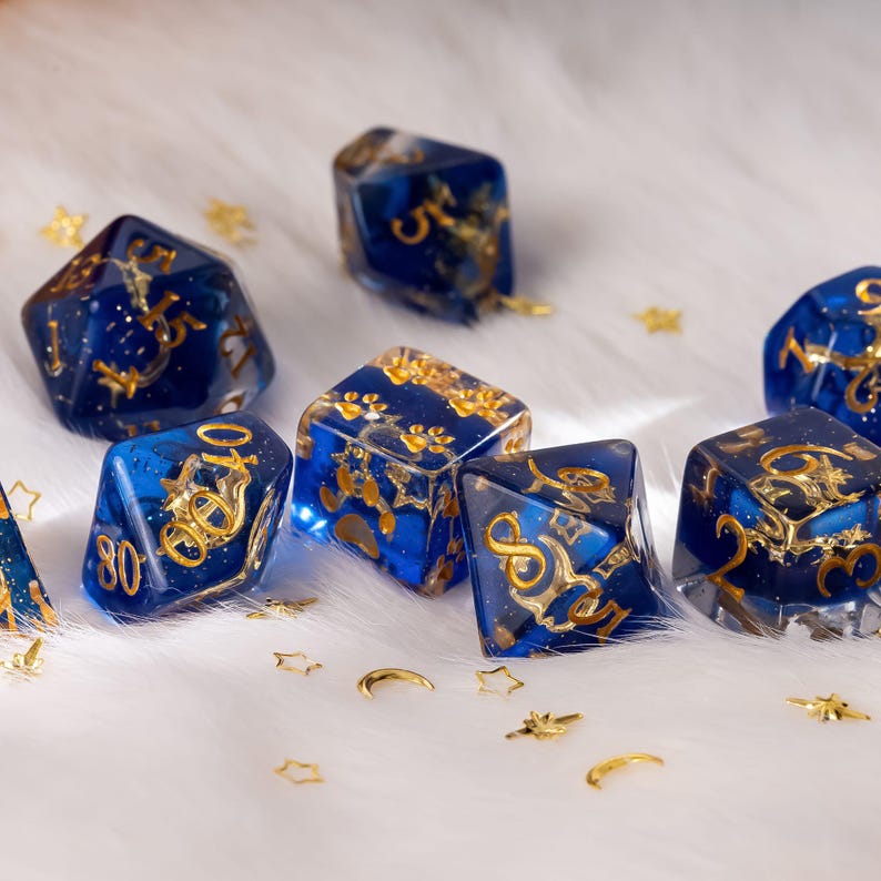 May include: A collection of blue and gold resin dice with gold numbering and accents. The dice are scattered on a white, fluffy surface, with small gold star and crescent moon decorations.