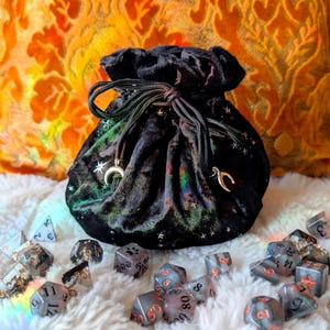 May include: A black velvet dice bag with a drawstring closure, decorated with stars and crescent moon charms. The bag is surrounded by polyhedral dice in grey, black, and clear, ideal for tabletop gaming.