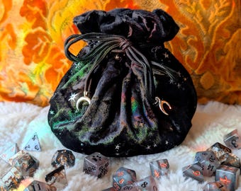 May include: Black velvet dice bag with a drawstring closure, adorned with celestial stars and crescent moon charms. The bag is surrounded by various polyhedral dice in shades of gray, black, and clear, perfect for tabletop gaming.