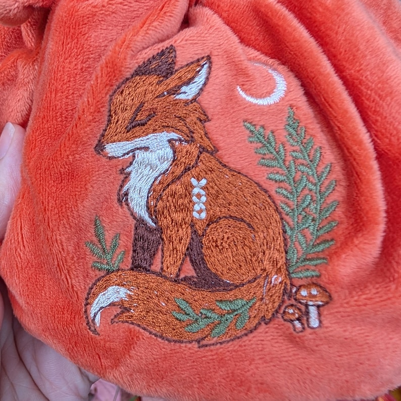 May include: An orange plush bag with an embroidered fox design. The fox is brown and white, with a crescent moon and foliage. The bag is soft and has a textured surface.