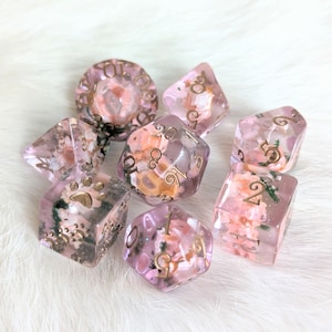 May include: Set of eight pink and gold dice with floral and swirl designs. The dice are translucent and have gold numbers.