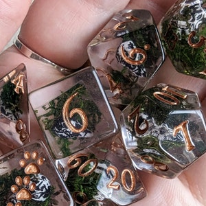 May include: A set of clear resin dice with gold-colored numbers and embedded green moss and small black and white details. The dice are in various polyhedral shapes, including a d20, d6, and d4, suitable for tabletop gaming.
