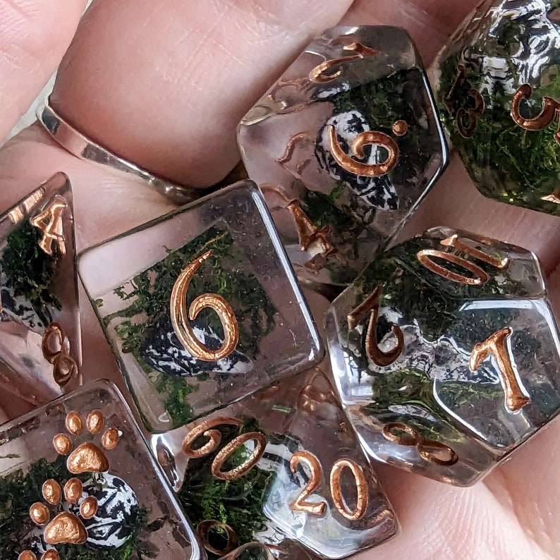May include: A set of clear resin dice with gold-colored numbers and embedded green moss and small black and white details. The dice are in various polyhedral shapes, including a d20, d6, and d4, suitable for tabletop gaming.