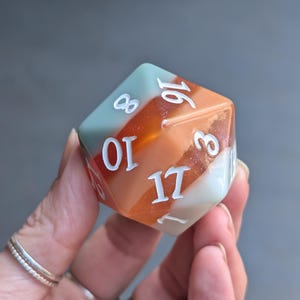May include: A twenty-sided die with white numerals, featuring sections of teal, orange, and white. The die is held in a hand, highlighting its design. Suitable for tabletop gaming. The die is a standard size for role-playing games.