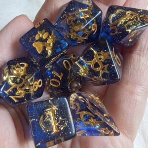 May include: A set of seven blue and gold dice with various symbols and numbers. The dice are made of a clear material with gold glitter embedded in it.
