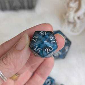 May include: A close-up of a twenty-sided die with a dark blue and black marbled design. The numbers are in a silver font. The die is held in a hand, with other dice visible in the background.
