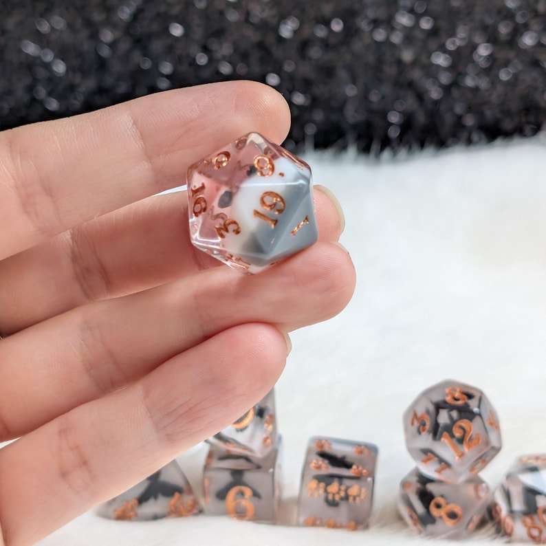 K&ouml;nnte beinhalten: Ein Satz polyedrischer W&uuml;rfel, darunter ein zwanzigseitiger W&uuml;rfel, mit einem transparenten, grauen und rot get&ouml;nten Design. Die Zahlen sind kupferfarben. Die W&uuml;rfel sind f&uuml;r Tabletop-Rollenspiele konzipiert.
