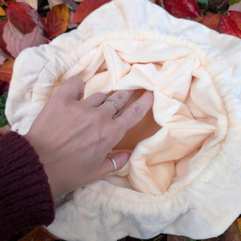 May include: A cream-colored, plush drawstring bag with a soft, quilted texture. The interior is a lighter shade of cream. The bag is open, revealing the inside. The image is taken from a top-down perspective.