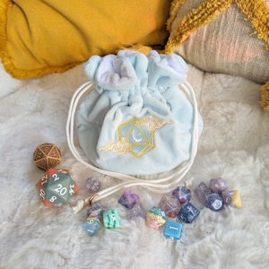 以下が含まれることがあります： 白い紐と金色の刺繍で月とサイコロのデザインが施された、薄い青色のベルベット製のサイコロ袋。袋にはカラフルなサイコロが入っています。