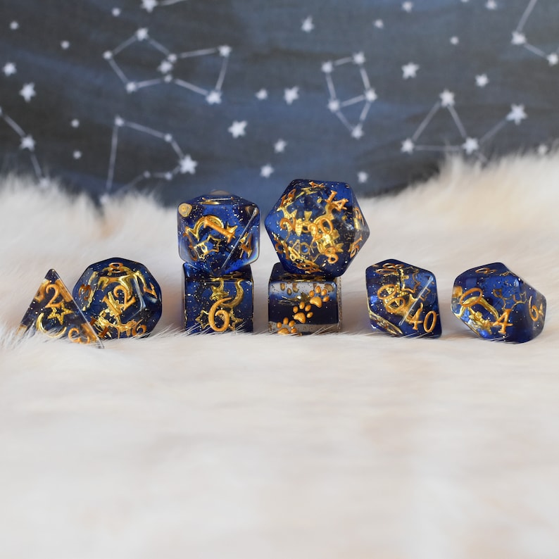 May include: Set of seven blue and gold dice with various constellations and symbols. The dice are made of resin and have a glittery finish.