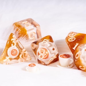 May include: A set of polyhedral dice made of translucent amber resin with embedded miniature white coffee cups featuring latte art. The dice have cream-coloured numbers and include a four-sided, eight-sided, and twenty-sided die, alongside two tiny loose coffee cup figurines.