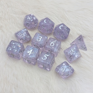 May include: A set of 10 purple dice with silver glitter and silver numbers. The dice are shaped like cubes and a pyramid. The dice are on a white fluffy surface.