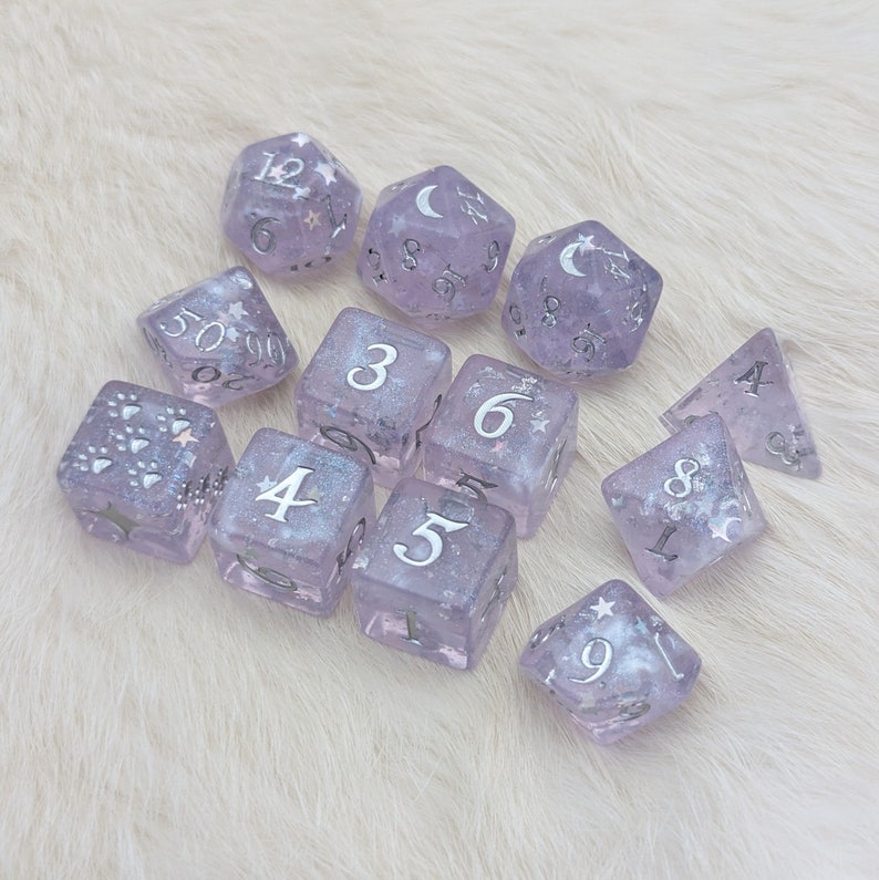 May include: A set of 10 purple dice with silver glitter and silver numbers. The dice are shaped like cubes and a pyramid. The dice are on a white fluffy surface.