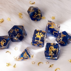 May include: A collection of blue and gold resin dice with gold numbering and decorative accents. The dice are scattered on a white, fluffy surface, with small gold stars and crescent moon shapes scattered around them. One die features a paw print.