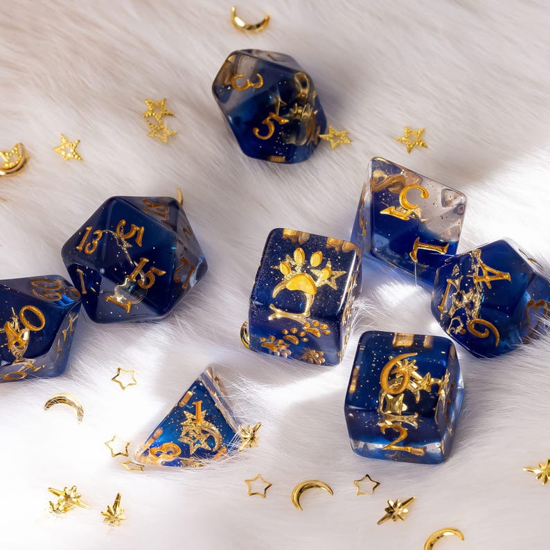 May include: A collection of blue and gold resin dice with gold numbering and decorative accents. The dice are scattered on a white, fluffy surface, with small gold stars and crescent moon shapes scattered around them. One die features a paw print.