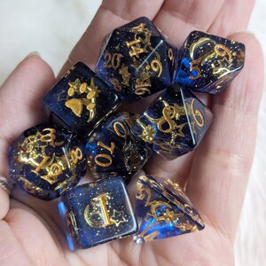 May include: Set of seven blue and gold dice with various symbols and numbers. The dice are made of clear blue resin with gold glitter and gold foil accents.