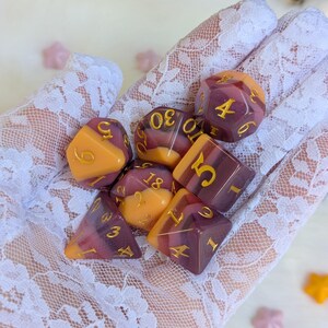 May include: A set of seven dice with a purple and orange swirl pattern. The dice have gold numbers on them. The dice are on a white lace surface.