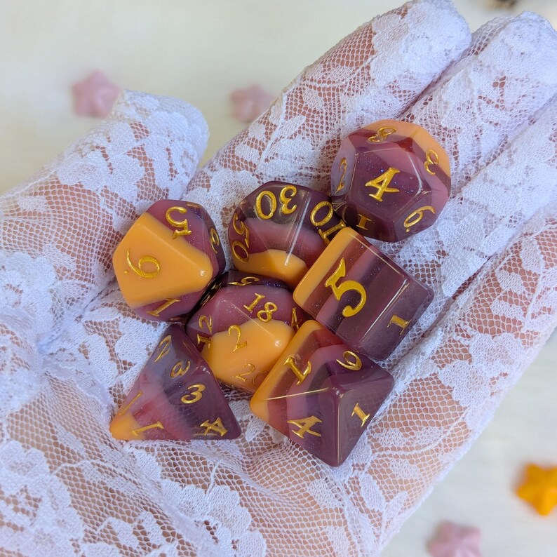May include: A set of seven dice with a purple and orange swirl pattern. The dice have gold numbers on them. The dice are on a white lace surface.