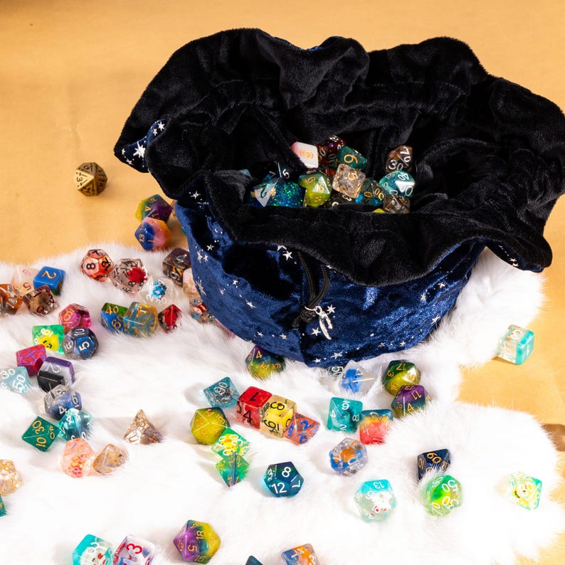 May include: A dark blue velvet drawstring pouch with a white star pattern and black interior lining, filled with a large collection of multi-colored, translucent polyhedral dice scattered across a white faux fur surface.