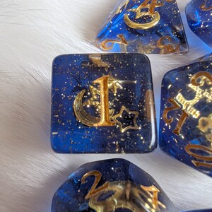 May include: A set of six blue dice with gold glitter and gold metallic accents. Each die has a different number from one to six, with a crescent moon and star design.