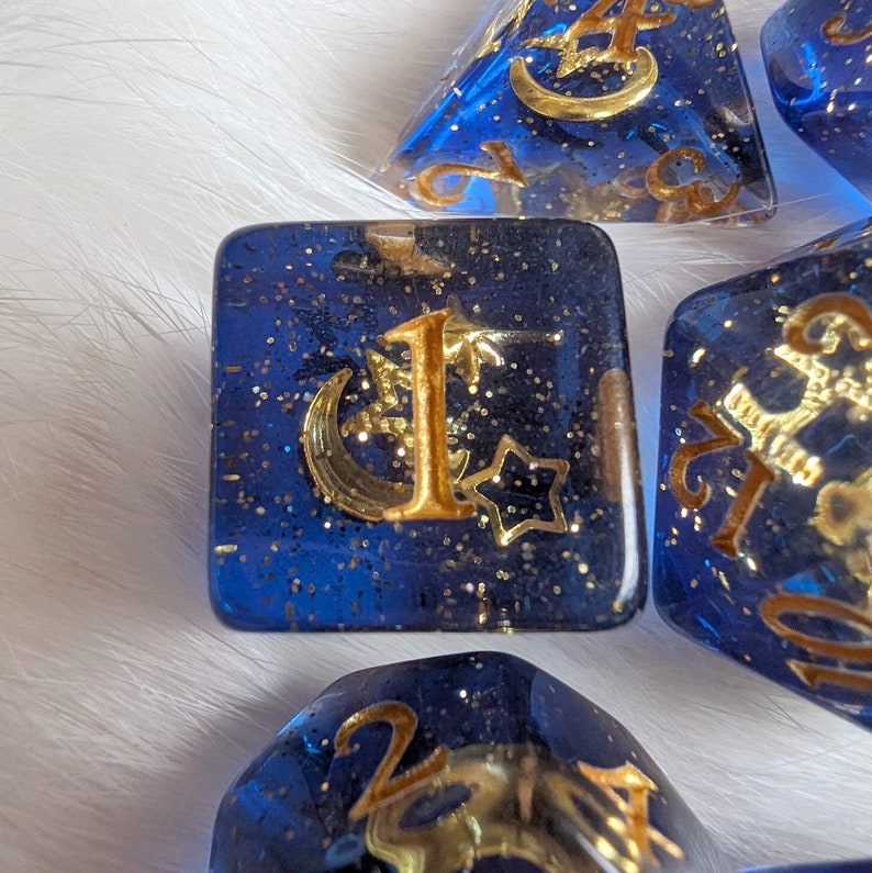 May include: A set of six blue dice with gold glitter and gold metallic accents. Each die has a different number from one to six, with a crescent moon and star design.