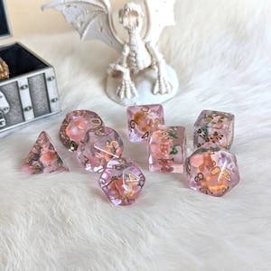 May include: A set of eight pink and gold dice with floral accents. The dice are made of clear resin with embedded flowers and gold glitter. The numbers on the dice are in gold.