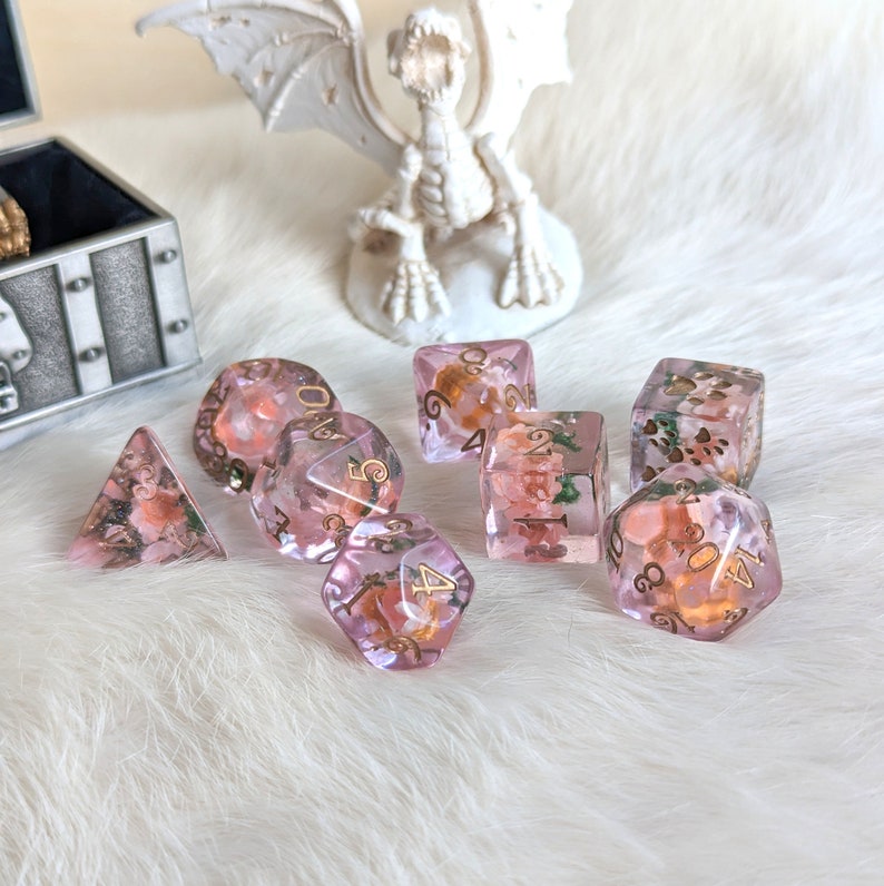 May include: A set of eight pink and gold dice with floral accents. The dice are made of clear resin with embedded flowers and gold glitter. The numbers on the dice are in gold.
